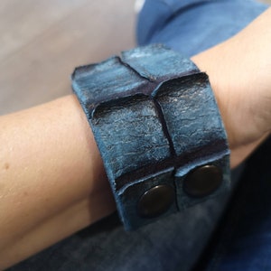 Puede incluir: Un brazalete de cuero azul con dos broches de metal negro. El brazalete est&aacute; hecho de cuero texturizado y tiene un aspecto desgastado.