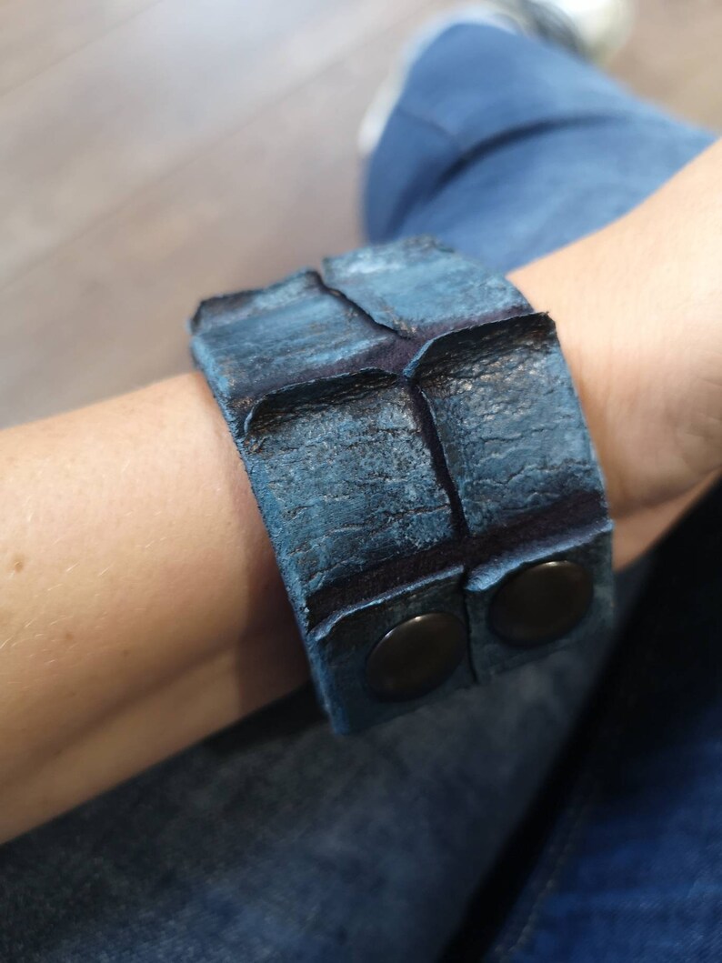 Puede incluir: Un brazalete de cuero azul con dos broches de metal negro. El brazalete est&aacute; hecho de cuero texturizado y tiene un aspecto desgastado.