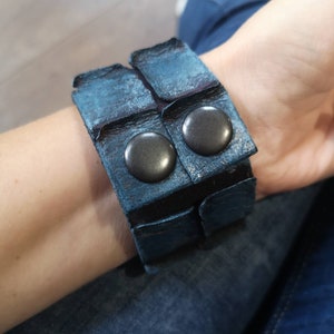Puede incluir: Un brazalete de cuero azul con dos tachuelas de metal negro. El brazalete est&aacute; hecho de varias capas de cuero y tiene un aspecto desgastado.