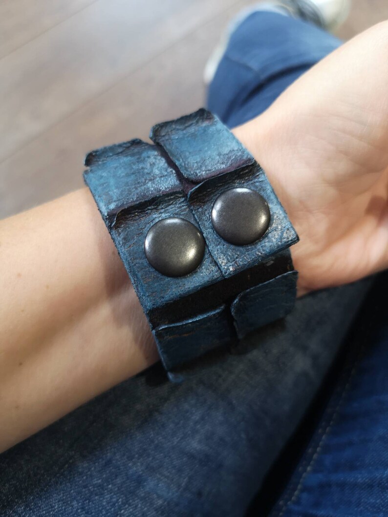 Puede incluir: Un brazalete de cuero azul con dos tachuelas de metal negro. El brazalete est&aacute; hecho de varias capas de cuero y tiene un aspecto desgastado.