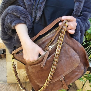 Peut inclure: Un sac &agrave; bandouli&egrave;re en cuir marron avec une cha&icirc;ne dor&eacute;e. Le sac a une poche avant et un compartiment principal zipp&eacute;. Le sac est tenu par une personne portant une veste en fausse fourrure bleu fonc&eacute;.