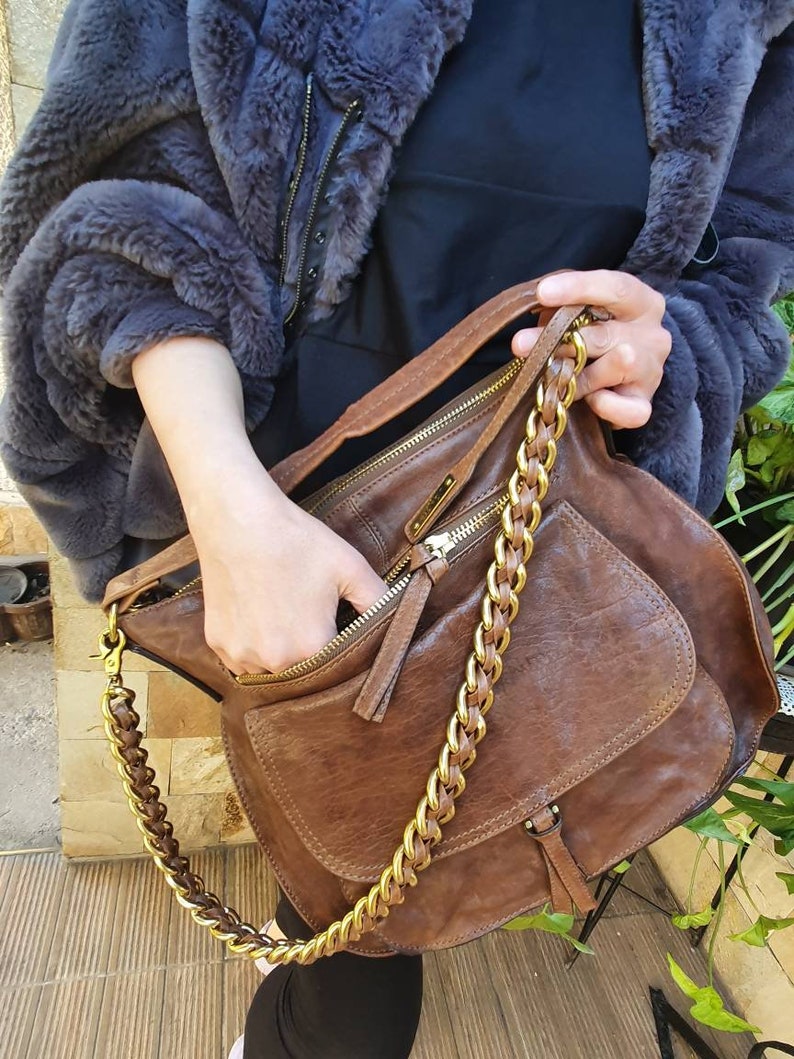 Peut inclure: Un sac &agrave; bandouli&egrave;re en cuir marron avec une cha&icirc;ne dor&eacute;e. Le sac a une poche avant et un compartiment principal zipp&eacute;. Le sac est tenu par une personne portant une veste en fausse fourrure bleu fonc&eacute;.
