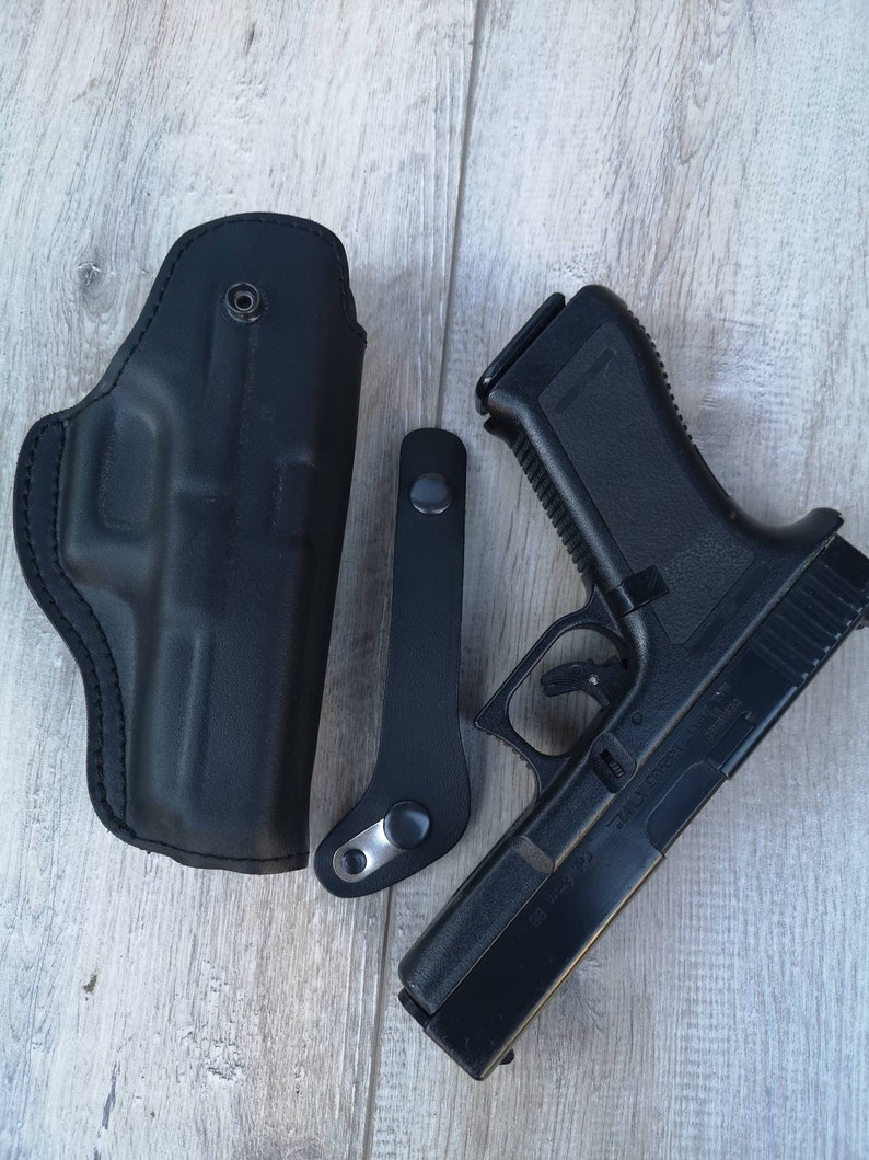 Puede incluir: Una funda de cuero negra con un clip de metal y una pistola negra con un marco de pol&iacute;mero negro.