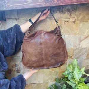 Peut inclure: Un sac &agrave; bandouli&egrave;re en cuir marron avec une seule sangle. Le sac a un design ample et un aspect us&eacute;. Le sac est tenu par les mains d'une personne. L'arri&egrave;re-plan est un mur de pierre.