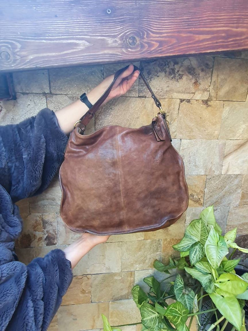 Peut inclure: Un sac &agrave; bandouli&egrave;re en cuir marron avec une seule sangle. Le sac a un design ample et un aspect us&eacute;. Le sac est tenu par les mains d'une personne. L'arri&egrave;re-plan est un mur de pierre.