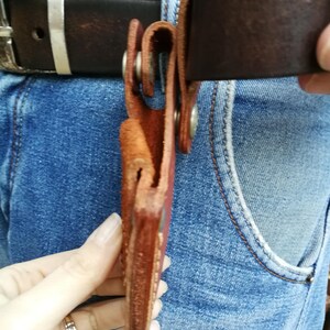 Pode incluir: Um coldre de couro marrom com fecho de press&atilde;o met&aacute;lico. O coldre est&aacute; preso a um cinto de couro marrom.