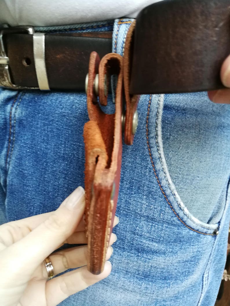 Pode incluir: Um coldre de couro marrom com fecho de press&atilde;o met&aacute;lico. O coldre est&aacute; preso a um cinto de couro marrom.