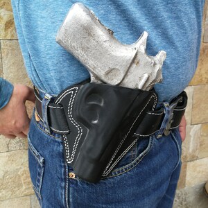 Puede incluir: Una pistola plateada en una funda de cuero negra con costuras blancas. La funda est&aacute; unida a un cintur&oacute;n de cuero marr&oacute;n que se lleva sobre unos vaqueros azules.