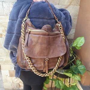 Peut inclure: Sac &agrave; bandouli&egrave;re en cuir marron avec une cha&icirc;ne dor&eacute;e. Le sac a une fermeture &agrave; rabat et une poche zipp&eacute;e. Le sac est port&eacute; sur un manteau de fourrure gris.