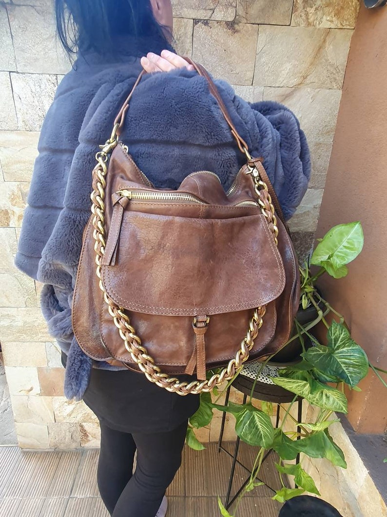 Peut inclure: Sac &agrave; bandouli&egrave;re en cuir marron avec une cha&icirc;ne dor&eacute;e. Le sac a une fermeture &agrave; rabat et une poche zipp&eacute;e. Le sac est port&eacute; sur un manteau de fourrure gris.