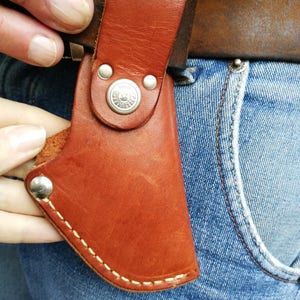 Pode incluir: Uma bainha de couro marrom com um bot&atilde;o prateado e costuras. A bainha est&aacute; presa a um cinto e &eacute; usada em um par de cal&ccedil;as jeans azuis.