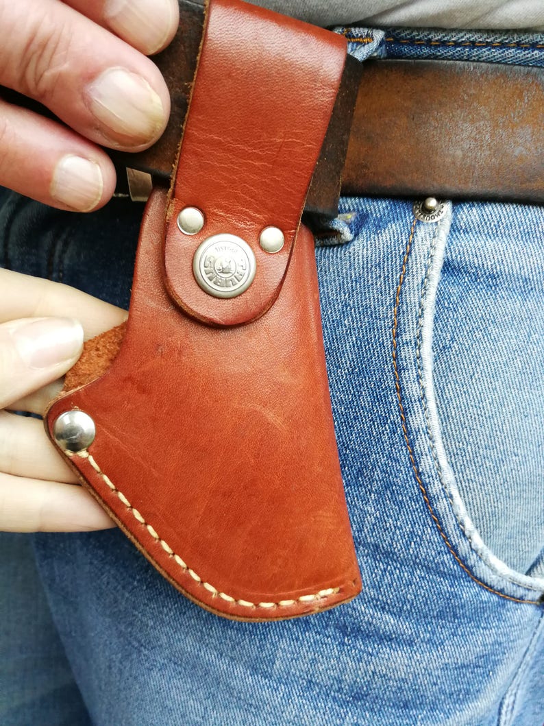 Pode incluir: Uma bainha de couro marrom com um bot&atilde;o prateado e costuras. A bainha est&aacute; presa a um cinto e &eacute; usada em um par de cal&ccedil;as jeans azuis.