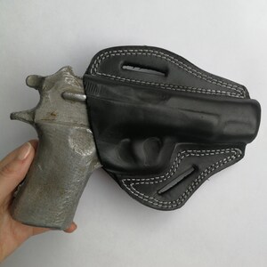 Puede incluir: Una funda de cuero negra con costuras blancas para una pistola. La funda tiene un bucle para el cintur&oacute;n y una traba para el pulgar.