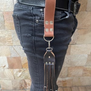 Könnte beinhalten: Eine braune Ledergürtelschlaufe mit einem schwarzen Lederquaste, die an einem silbernen Schlüsselring befestigt ist. Die Schlaufe ist an einem braunen Ledergürtel befestigt.