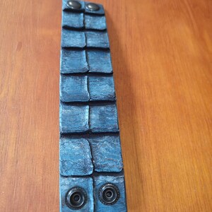 Puede incluir: Un brazalete de cuero azul con una superficie texturizada y dos broches met&aacute;licos negros.