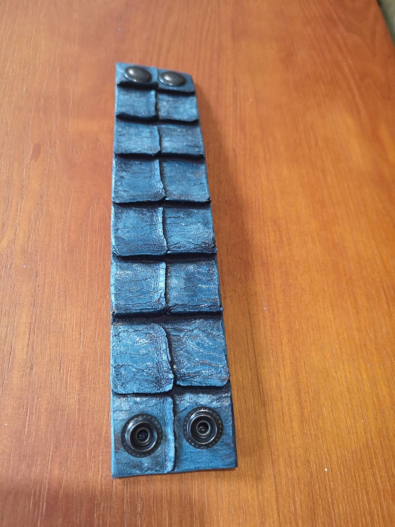 Puede incluir: Un brazalete de cuero azul con una superficie texturizada y dos broches met&aacute;licos negros.