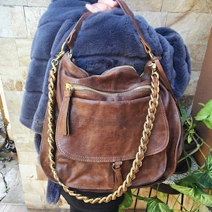 Peut inclure: Sac &agrave; bandouli&egrave;re en cuir marron avec une cha&icirc;ne dor&eacute;e. Le sac poss&egrave;de une poche &agrave; rabat &agrave; l'avant et une fermeture &eacute;clair sur le dessus. Le cuir a un aspect vieilli. Le sac est port&eacute; &agrave; l'&eacute;paule.
