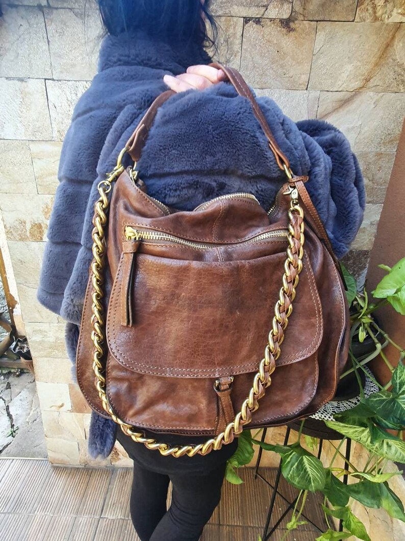 Peut inclure: Sac &agrave; bandouli&egrave;re en cuir marron avec une cha&icirc;ne dor&eacute;e. Le sac poss&egrave;de une poche &agrave; rabat &agrave; l'avant et une fermeture &eacute;clair sur le dessus. Le cuir a un aspect vieilli. Le sac est port&eacute; &agrave; l'&eacute;paule.