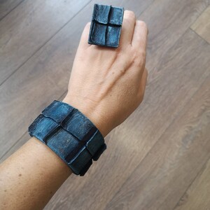 Puede incluir: Un conjunto de pulsera y anillo de cuero azul con un dise&ntilde;o geom&eacute;trico. La pulsera es ancha y tiene forma cuadrada. El anillo tambi&eacute;n tiene forma cuadrada y tiene un dise&ntilde;o similar al de la pulsera.