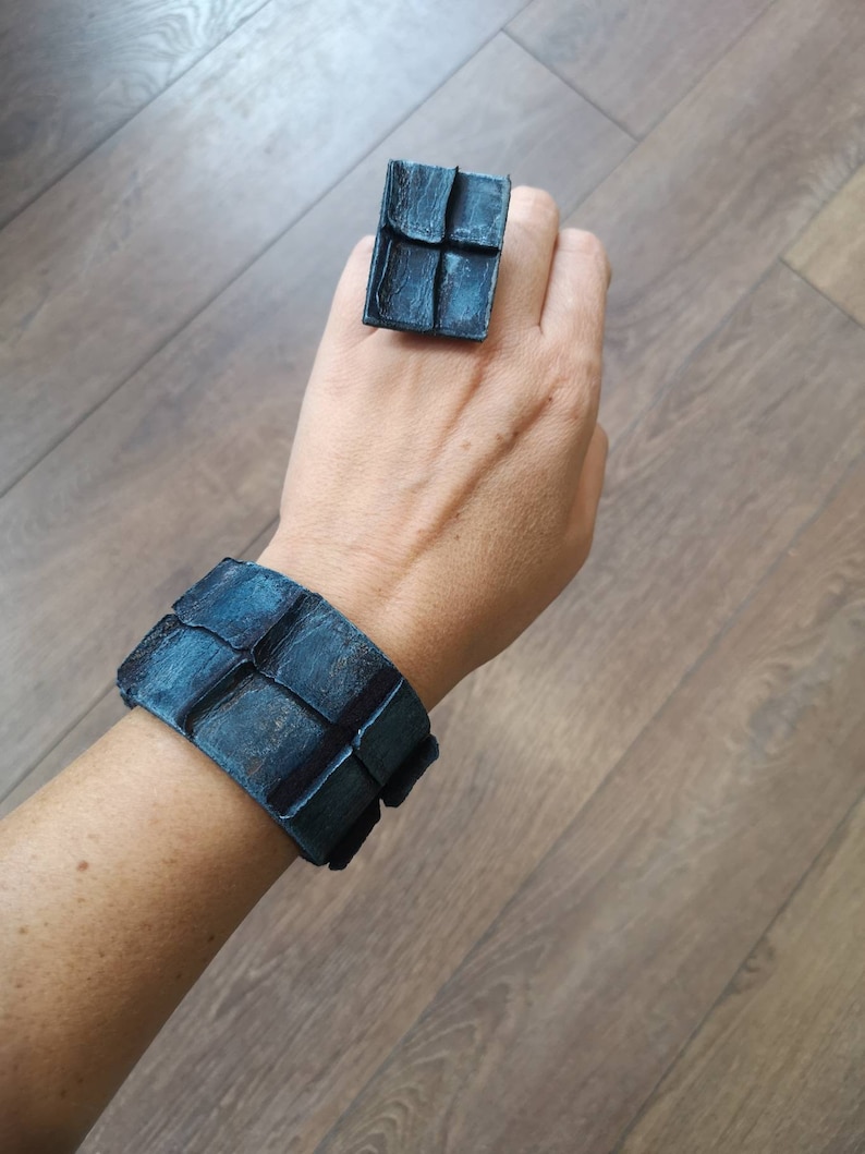 Puede incluir: Un conjunto de pulsera y anillo de cuero azul con un dise&ntilde;o geom&eacute;trico. La pulsera es ancha y tiene forma cuadrada. El anillo tambi&eacute;n tiene forma cuadrada y tiene un dise&ntilde;o similar al de la pulsera.