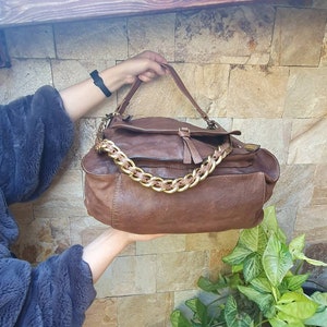 Peut inclure: Un sac &agrave; main en cuir marron avec une cha&icirc;ne dor&eacute;e. Le sac a une fermeture &agrave; rabat et un aspect textur&eacute;. Le sac est tenu par une personne. L'arri&egrave;re-plan est un mur de pierre et des plantes vertes.
