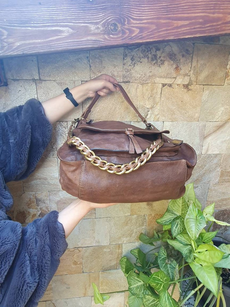 Peut inclure: Un sac &agrave; main en cuir marron avec une cha&icirc;ne dor&eacute;e. Le sac a une fermeture &agrave; rabat et un aspect textur&eacute;. Le sac est tenu par une personne. L'arri&egrave;re-plan est un mur de pierre et des plantes vertes.