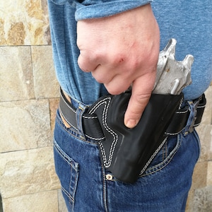 Puede incluir: Una funda de cuero negra con un dise&ntilde;o de puntadas blancas, que se lleva en la cadera derecha de una persona, con una pistola plateada en su interior.