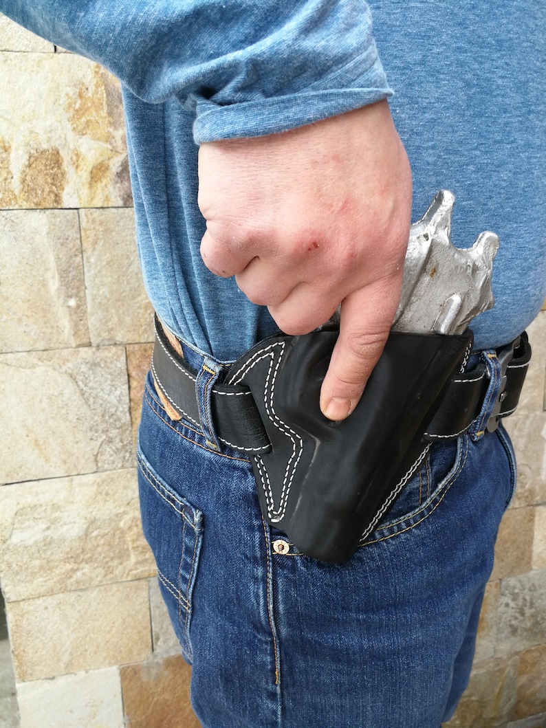 Puede incluir: Una funda de cuero negra con un dise&ntilde;o de puntadas blancas, que se lleva en la cadera derecha de una persona, con una pistola plateada en su interior.