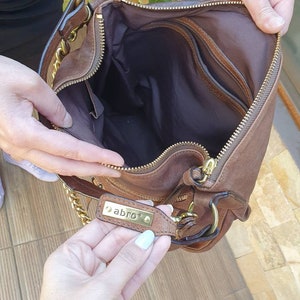 Peut inclure: Un sac &agrave; main en cuir marron avec une bandouli&egrave;re en cha&icirc;ne dor&eacute;e et une fermeture &eacute;clair. L'int&eacute;rieur est marron fonc&eacute; avec plusieurs poches. Le sac &agrave; main porte une &eacute;tiquette m&eacute;tallique dor&eacute;e avec le mot "abro".