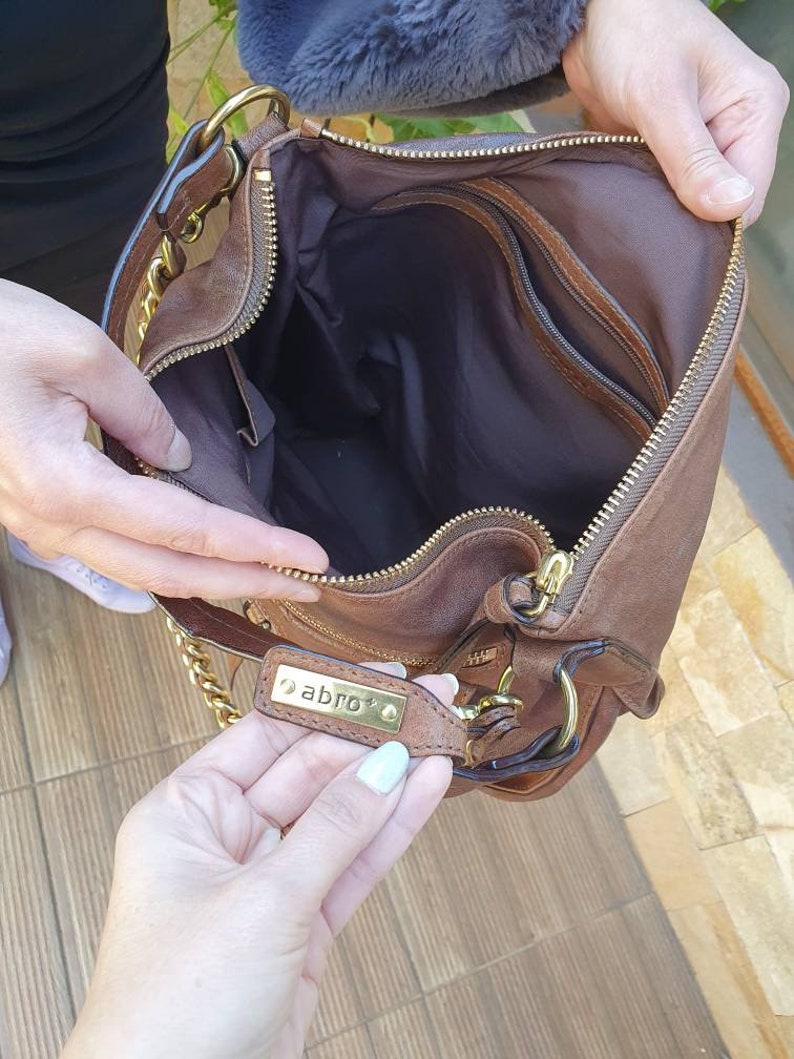Peut inclure: Un sac &agrave; main en cuir marron avec une bandouli&egrave;re en cha&icirc;ne dor&eacute;e et une fermeture &eacute;clair. L'int&eacute;rieur est marron fonc&eacute; avec plusieurs poches. Le sac &agrave; main porte une &eacute;tiquette m&eacute;tallique dor&eacute;e avec le mot "abro".