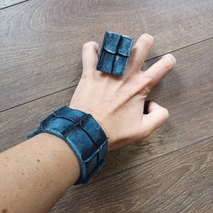 Puede incluir: Un conjunto de pulsera y anillo de cuero azul con un dise&ntilde;o geom&eacute;trico. La pulsera es ancha y tiene una superficie texturizada. El anillo es de forma cuadrada y tiene una superficie texturizada similar.