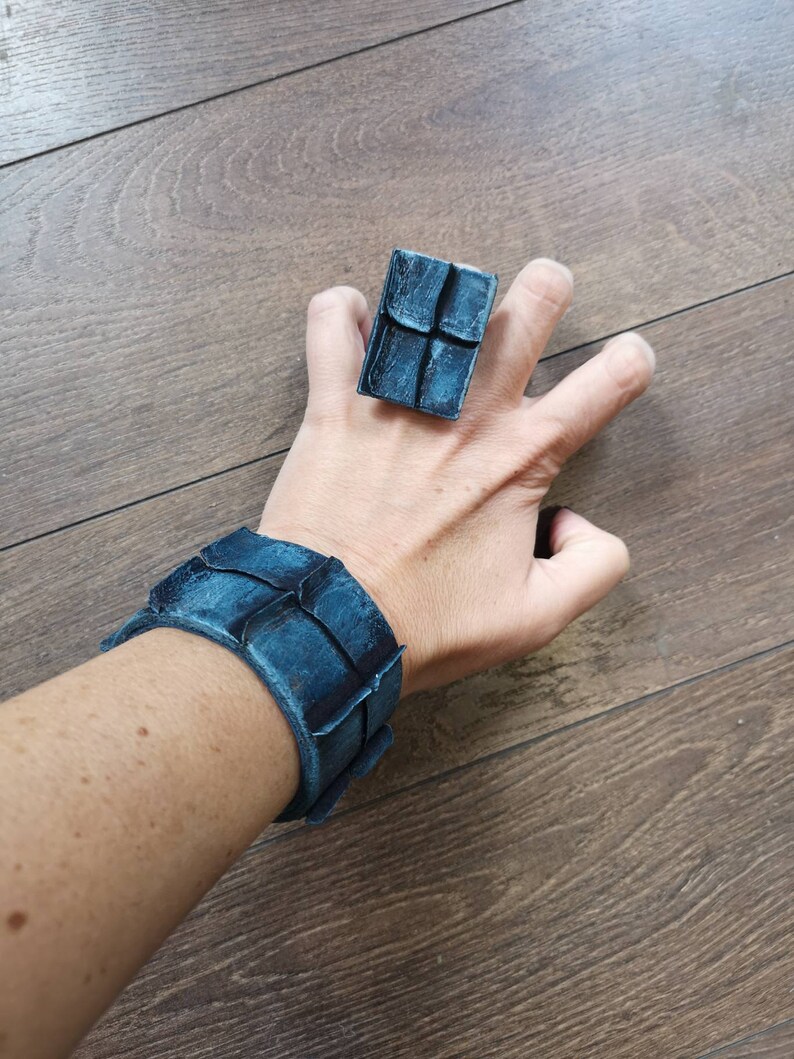 Puede incluir: Un conjunto de pulsera y anillo de cuero azul con un dise&ntilde;o geom&eacute;trico. La pulsera es ancha y tiene una superficie texturizada. El anillo es de forma cuadrada y tiene una superficie texturizada similar.