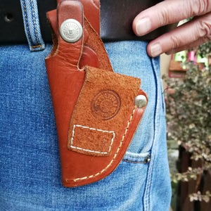 Pode incluir: Um coldre de couro marrom com um bot&atilde;o de metal e costuras. O coldre &eacute; preso a um cinto e &eacute; usado no quadril de uma pessoa.