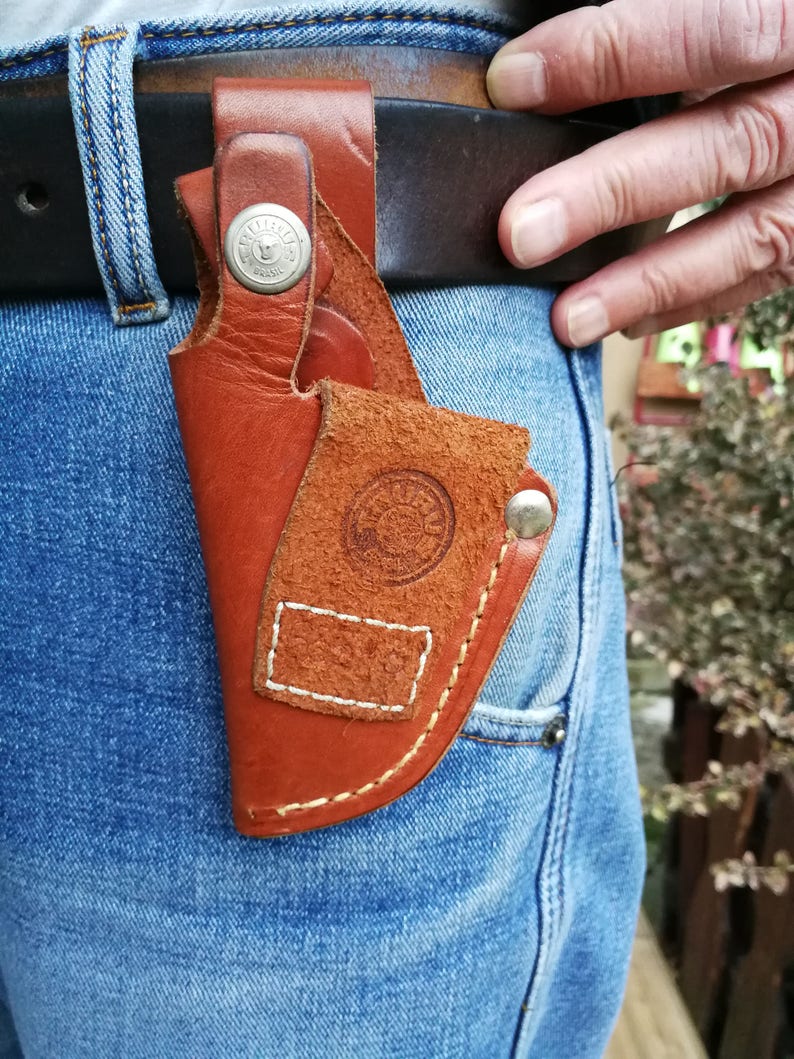 Pode incluir: Um coldre de couro marrom com um bot&atilde;o de metal e costuras. O coldre &eacute; preso a um cinto e &eacute; usado no quadril de uma pessoa.