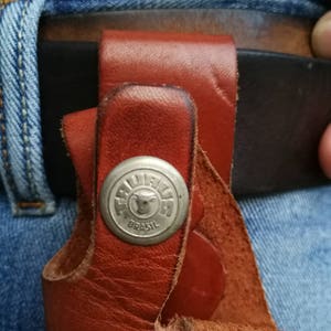 Pode incluir: Um coldre de couro marrom com um bot&atilde;o de metal prateado com um logotipo de cabe&ccedil;a de touro e o texto "TRULLI  BRASIL".