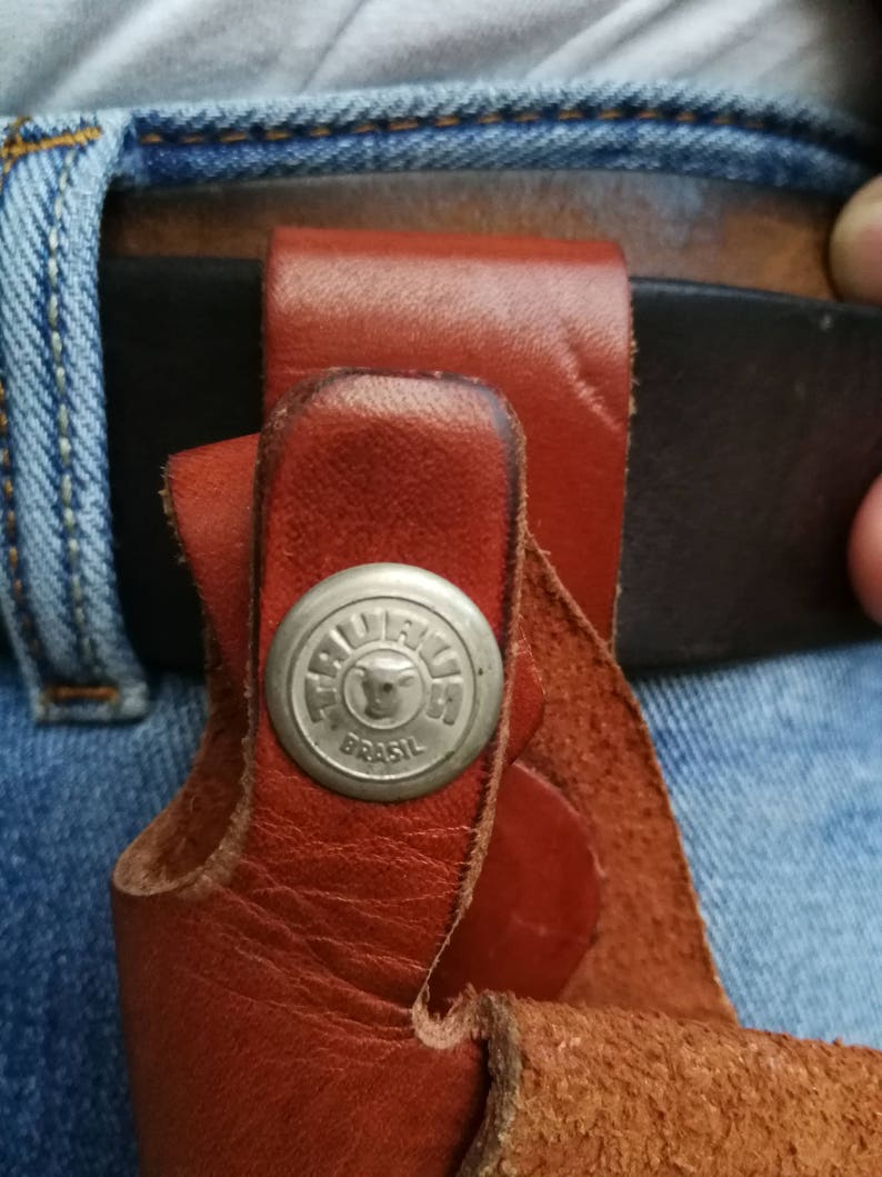 Pode incluir: Um coldre de couro marrom com um bot&atilde;o de metal prateado com um logotipo de cabe&ccedil;a de touro e o texto "TRULLI  BRASIL".