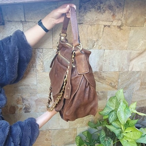 Peut inclure: Un sac &agrave; main en cuir marron avec des d&eacute;tails dor&eacute;s, dont une cha&icirc;ne et des fermetures &eacute;clair. Le sac a un design souple et une poign&eacute;e sup&eacute;rieure. Le fond pr&eacute;sente un mur en pierre et de la verdure.