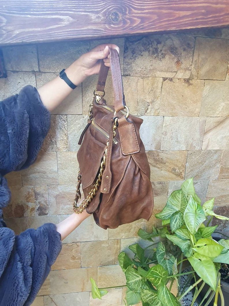 Peut inclure: Un sac &agrave; main en cuir marron avec des d&eacute;tails dor&eacute;s, dont une cha&icirc;ne et des fermetures &eacute;clair. Le sac a un design souple et une poign&eacute;e sup&eacute;rieure. Le fond pr&eacute;sente un mur en pierre et de la verdure.