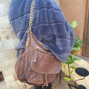 Peut inclure: Un sac &agrave; bandouli&egrave;re en cuir marron avec une cha&icirc;ne dor&eacute;e. Le sac a une poche &agrave; rabat et une poche zipp&eacute;e. La personne porte un manteau en fausse fourrure bleu.
