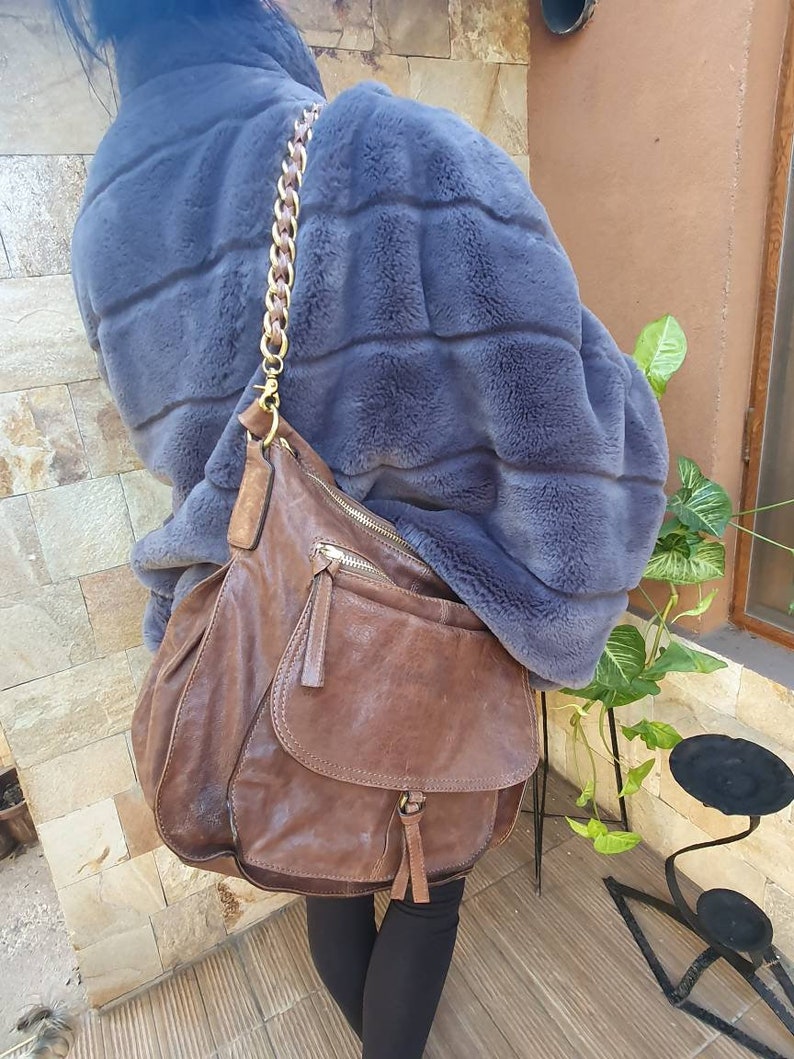 Peut inclure: Un sac &agrave; bandouli&egrave;re en cuir marron avec une cha&icirc;ne dor&eacute;e. Le sac a une poche &agrave; rabat et une poche zipp&eacute;e. La personne porte un manteau en fausse fourrure bleu.