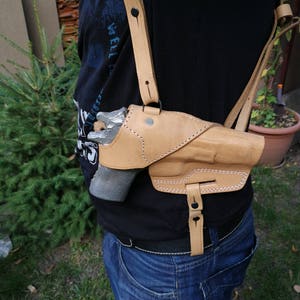 Puede incluir: Una funda de cuero marrón con una correa para el hombro y un botón a presión negro. La funda está diseñada para sostener una pistola.