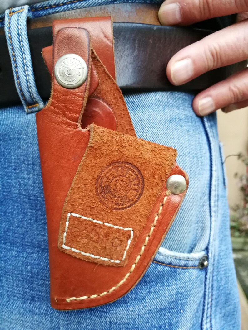 Pode incluir: Um coldre de couro marrom com detalhes de costura branca e um bot&atilde;o prateado. O coldre tem um logotipo circular com a palavra "BRASIL" impressa nele.