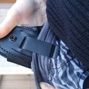 Puede incluir: Una funda de cuero negra con un clip met&aacute;lico sujeto a un pantal&oacute;n de rayas blancas y negras.