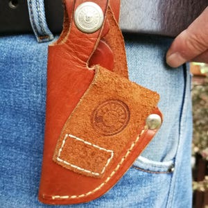 Pode incluir: Um coldre de couro marrom com um bot&atilde;o de metal e um design costurado. O coldre est&aacute; preso a um cinto.
