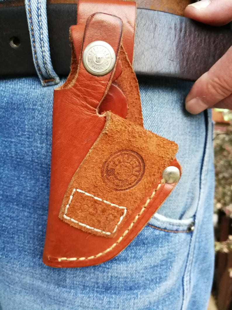 Pode incluir: Um coldre de couro marrom com um bot&atilde;o de metal e um design costurado. O coldre est&aacute; preso a um cinto.