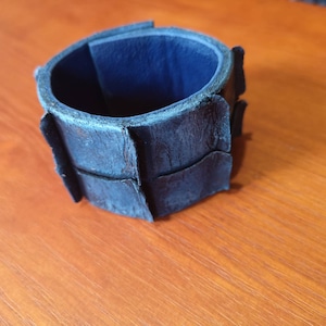 Puede incluir: Un brazalete de cuero azul oscuro con un dise&ntilde;o &uacute;nico segmentado. El brazalete est&aacute; hecho de m&uacute;ltiples piezas de cuero que se han cosido juntas para crear un aspecto texturizado.