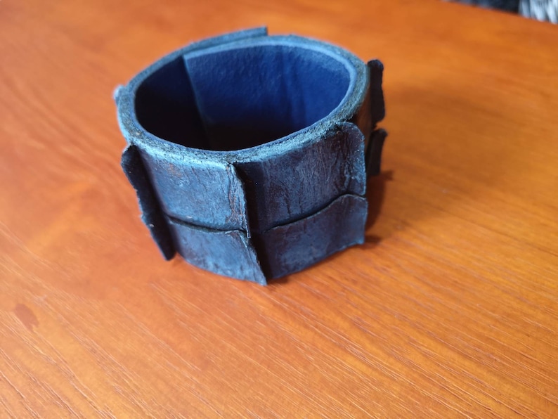 Puede incluir: Un brazalete de cuero azul oscuro con un dise&ntilde;o &uacute;nico segmentado. El brazalete est&aacute; hecho de m&uacute;ltiples piezas de cuero que se han cosido juntas para crear un aspecto texturizado.