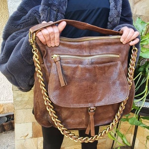 Peut inclure: Un sac &agrave; bandouli&egrave;re en cuir marron avec une cha&icirc;ne dor&eacute;e. Le sac a une fermeture &agrave; rabat et plusieurs compartiments zipp&eacute;s. Le sac est tenu par une personne portant un manteau en fausse fourrure grise.