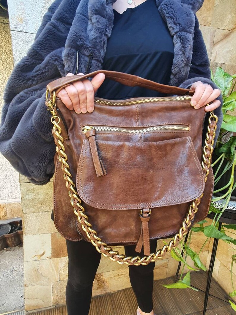 Peut inclure: Un sac &agrave; bandouli&egrave;re en cuir marron avec une cha&icirc;ne dor&eacute;e. Le sac a une fermeture &agrave; rabat et plusieurs compartiments zipp&eacute;s. Le sac est tenu par une personne portant un manteau en fausse fourrure grise.