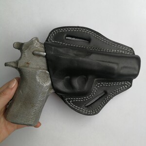 Puede incluir: Una funda de cuero negra con costuras blancas para una pistola. La funda est&aacute; dise&ntilde;ada para ajustarse perfectamente al arma y tiene una solapa que cubre el gatillo.